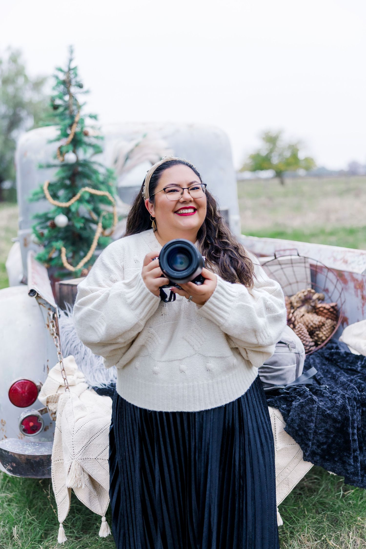 Smiling woman in cream sweater holding camera outdoors near vintage truck with Christmas tree.