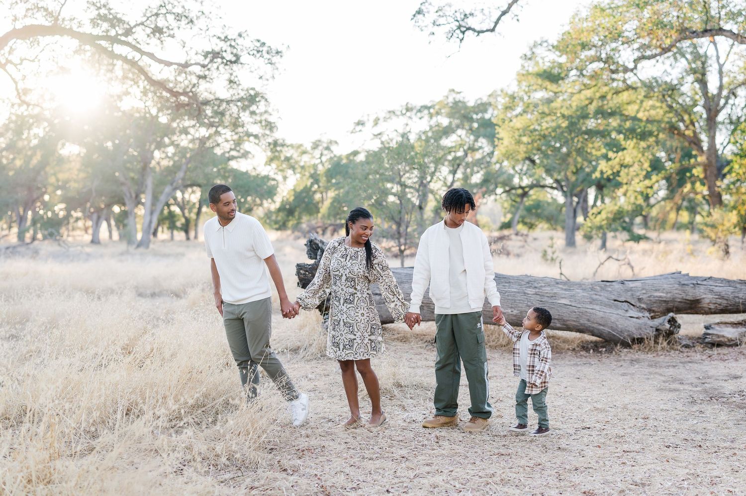 Family of five walking hand-in-hand outdoors in a sunlit field surrounded by trees during golden hour.