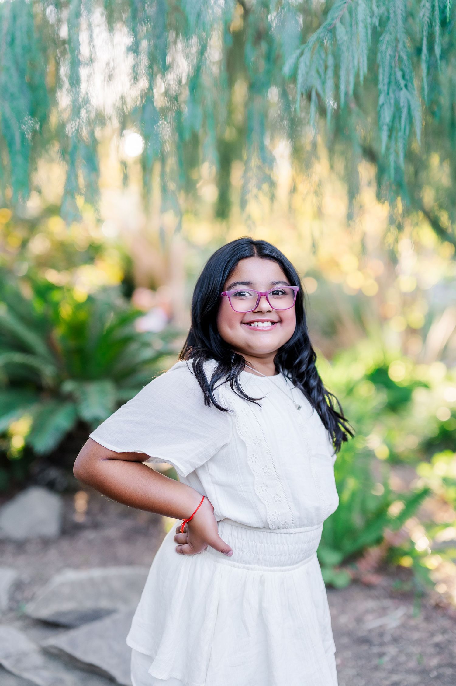 A young person in a white dress poses playfully outdoors with natural greenery in the background.