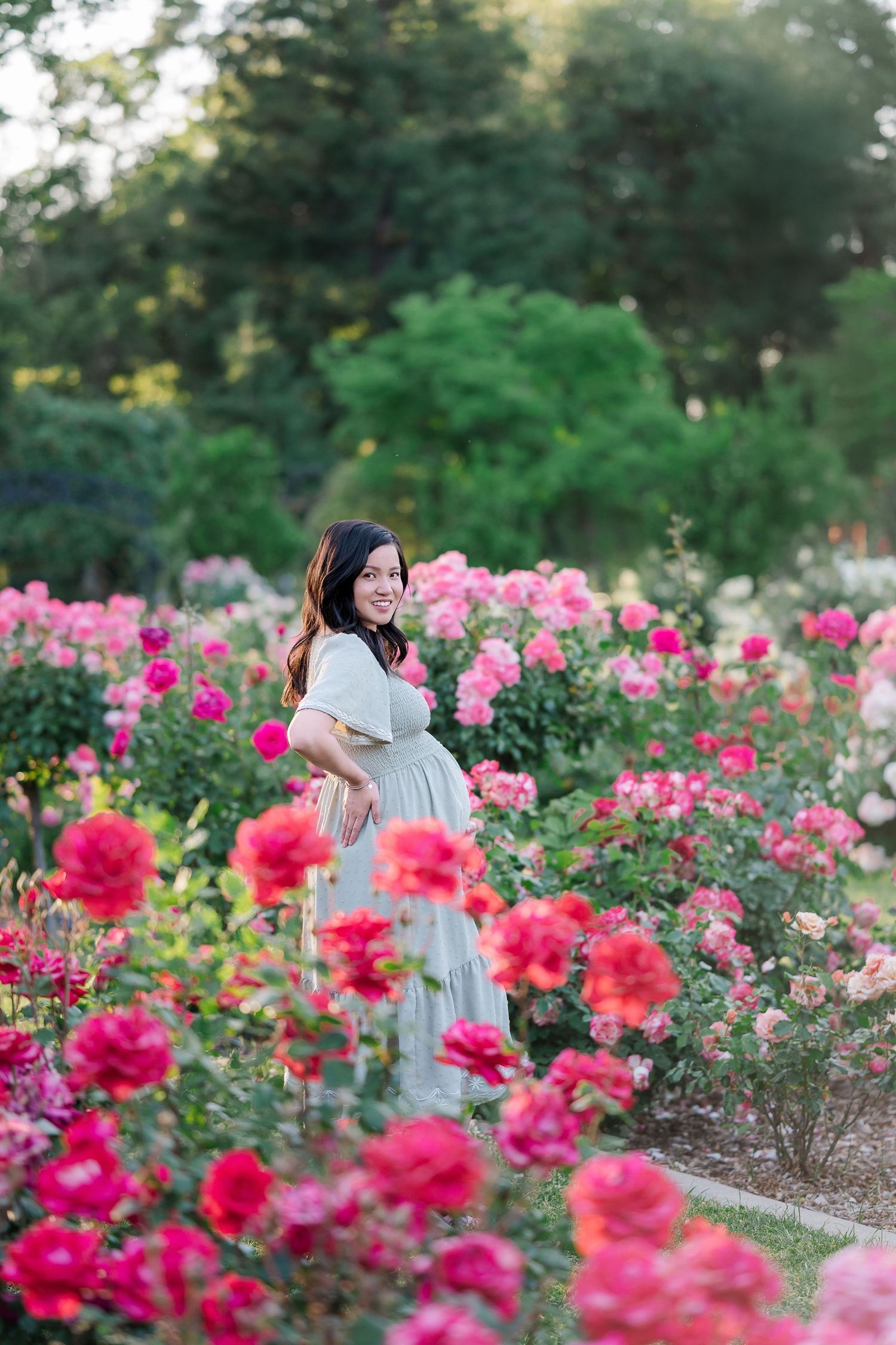 A person in a white dress explores a vibrant rose garden filled with pink and red blooming flowers on a summer day.