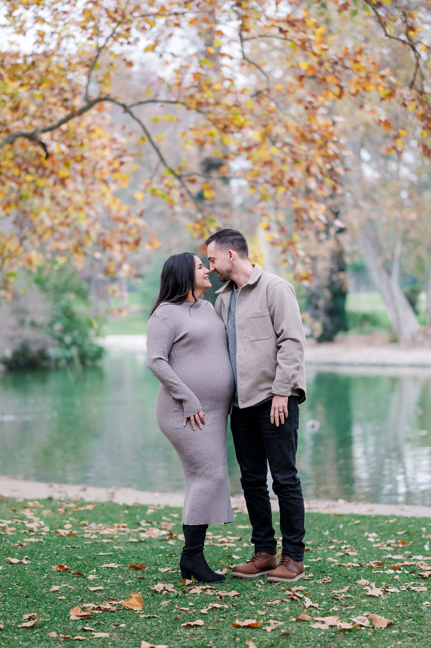 A couple shares a tender moment on an autumn day by a lake with golden leaves scattered on the ground.