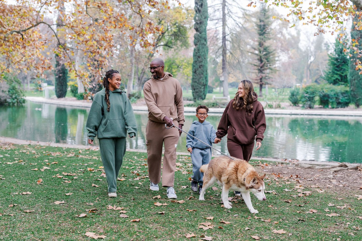 A group of people and a dog enjoy an autumn walk together through a scenic park with fallen leaves and a pond.