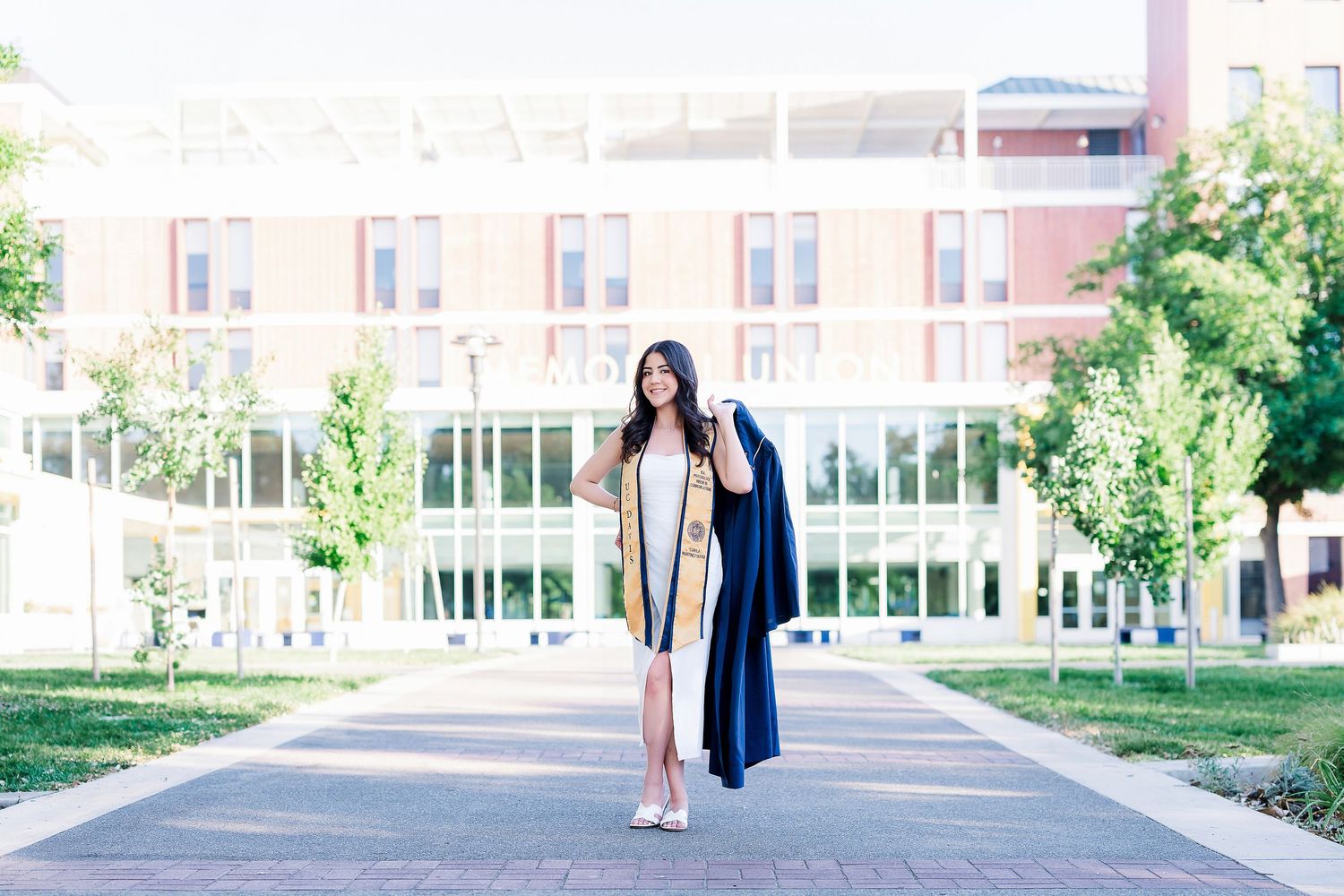 Graduate in cap and gown poses on campus pathway with brick building and trees in background.