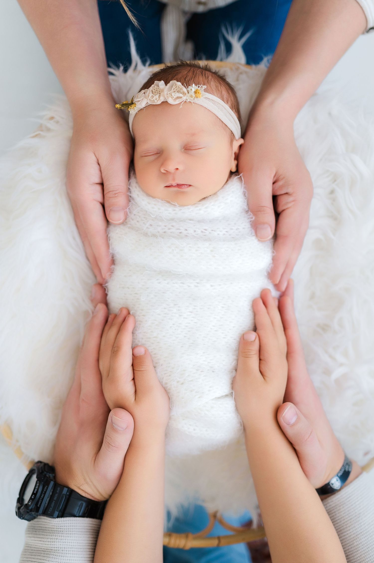 Newborn baby swaddled in white wrap with floral headband, cradled gently by parents hands on fluffy white surface.