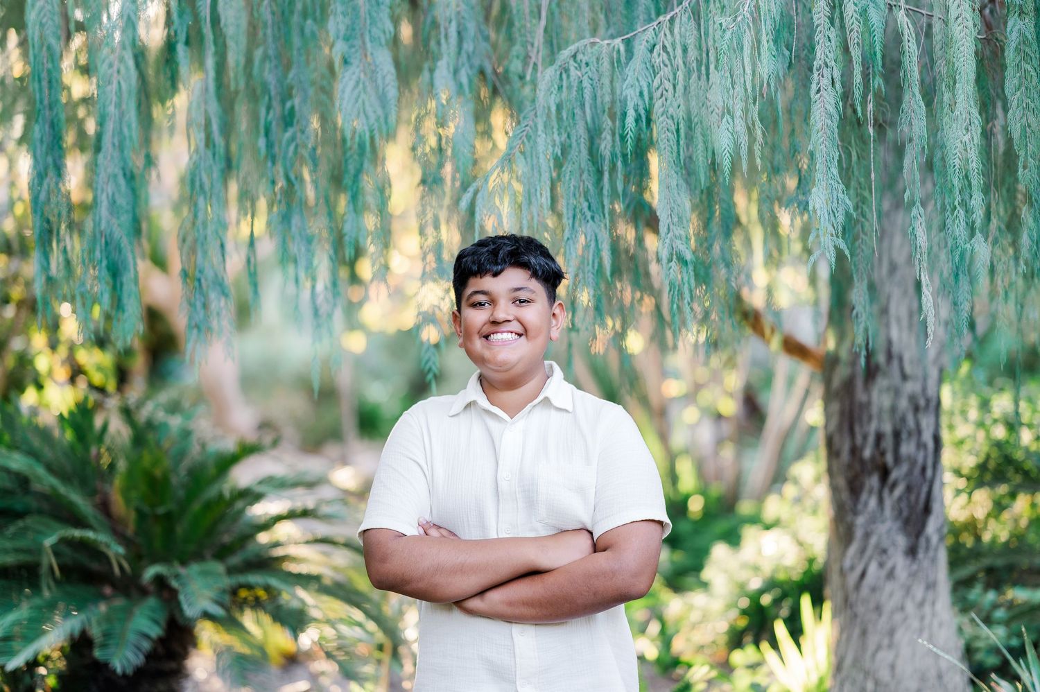 A person in a white shirt poses against a lush green garden background with palm trees and natural foliage.