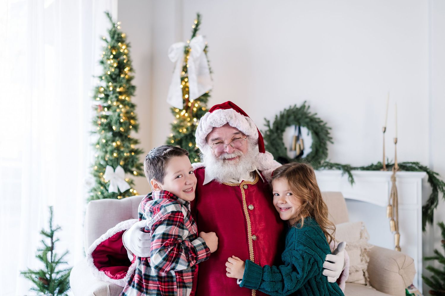Santa Claus hugs two smiling children in festive attire near a decorated Christmas tree and wreath indoors.
