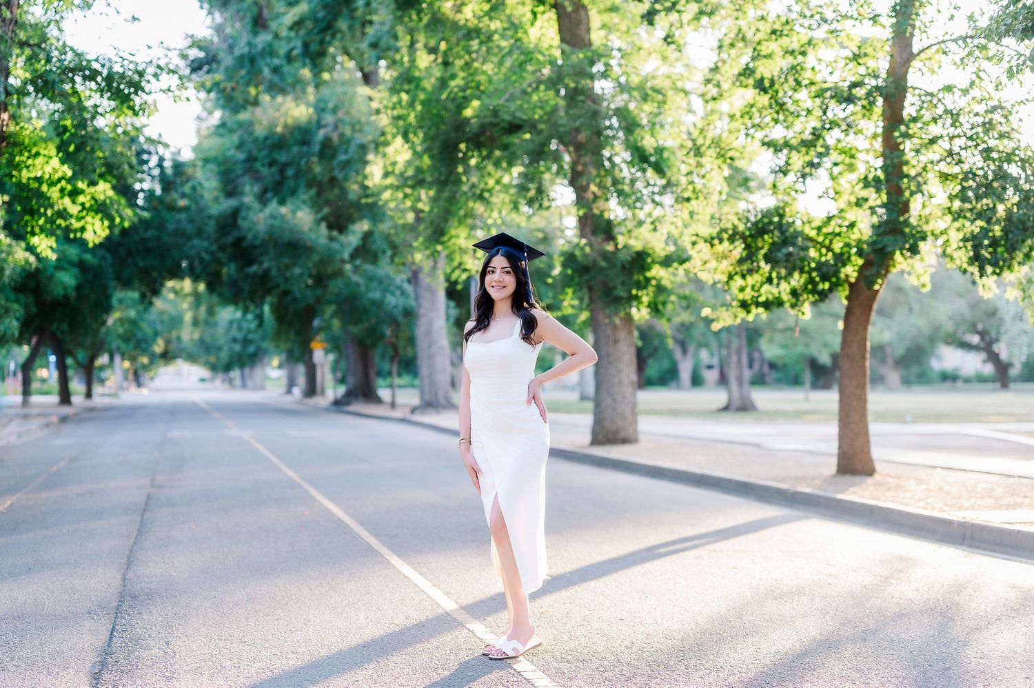 A graduate in white dress poses on a tree-lined path during golden hour sunlight.