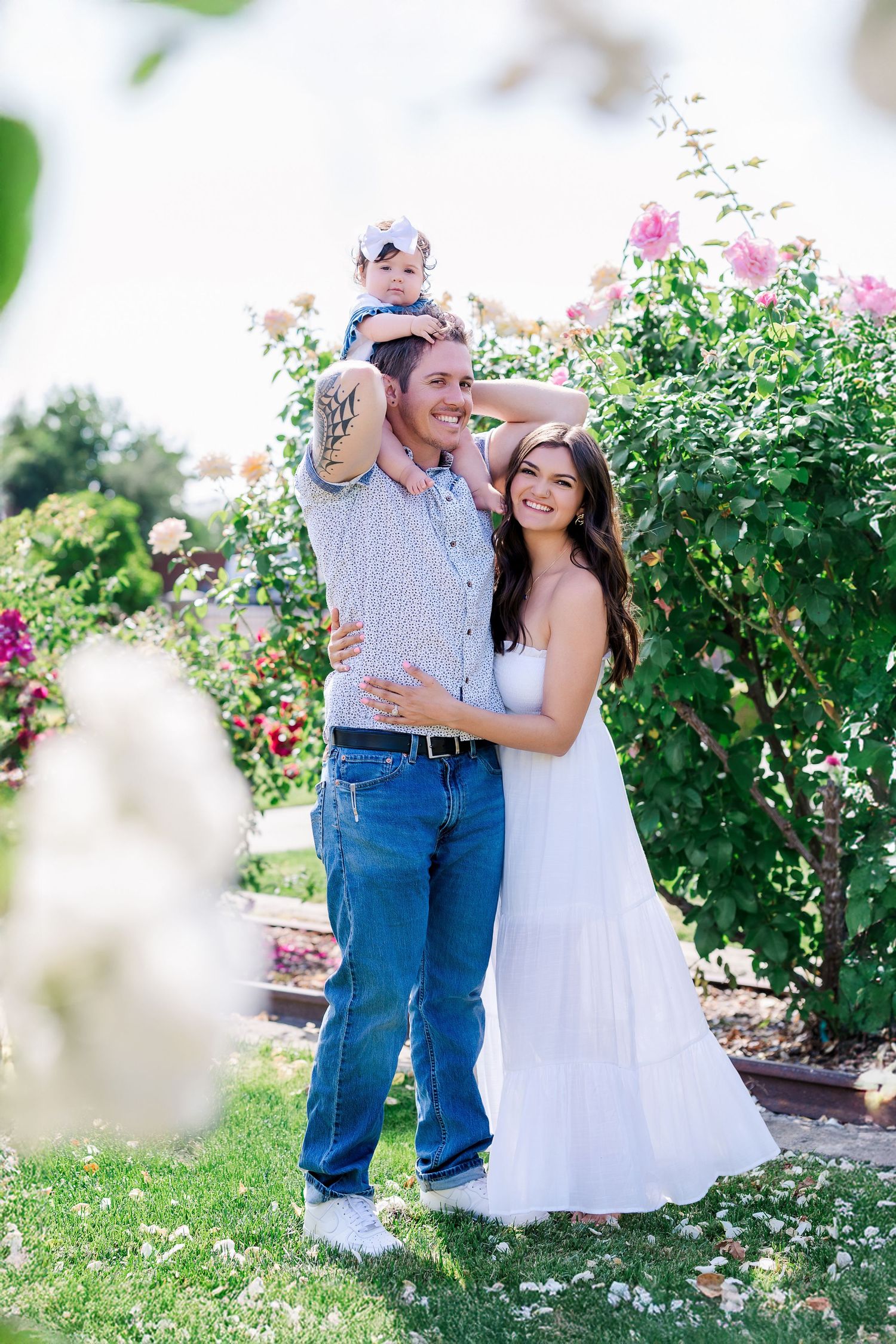 A couple embraces in a sunlit rose garden while holding their small child.