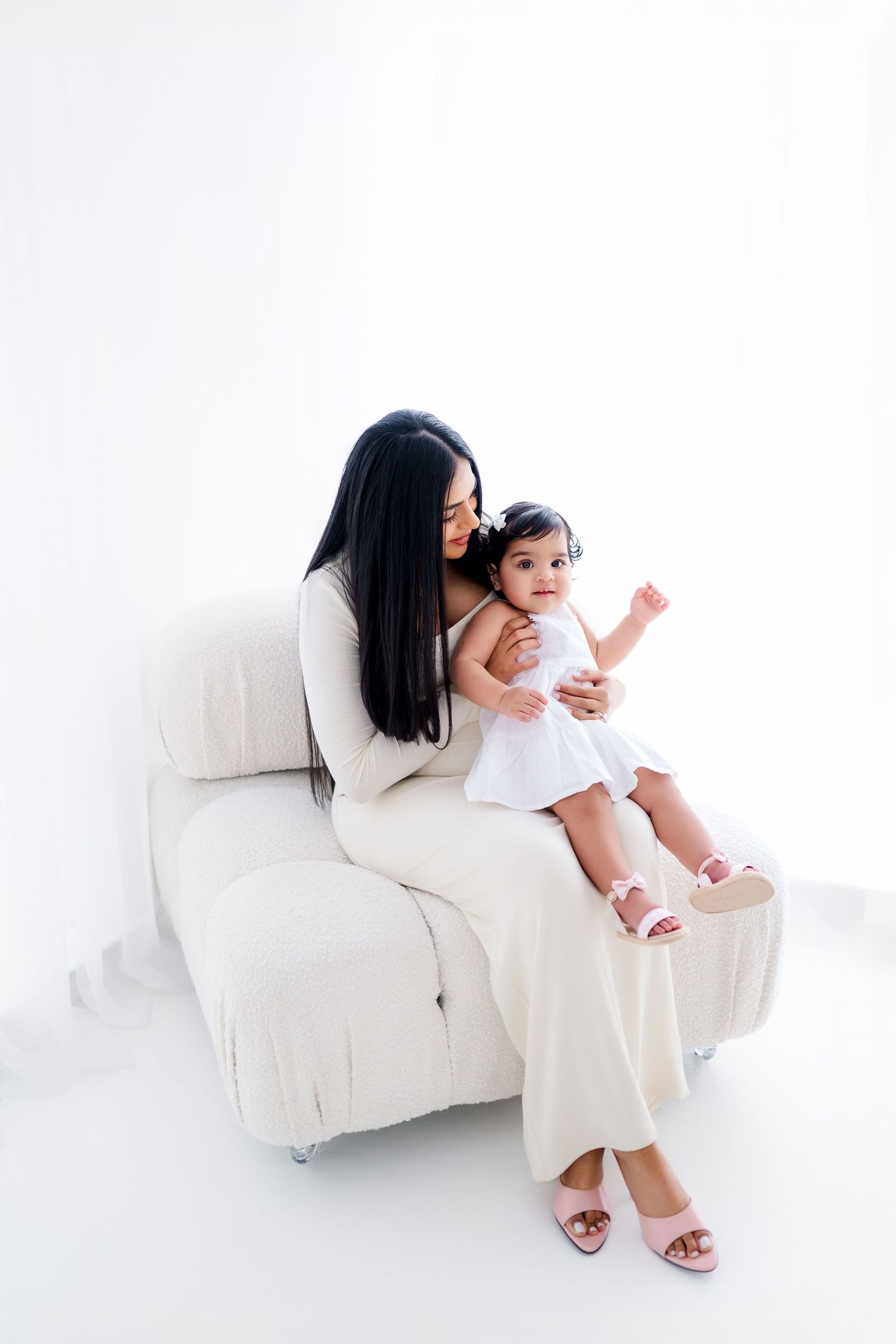 A parent and child wearing white clothing sit together on a white couch in a minimalist setting.
