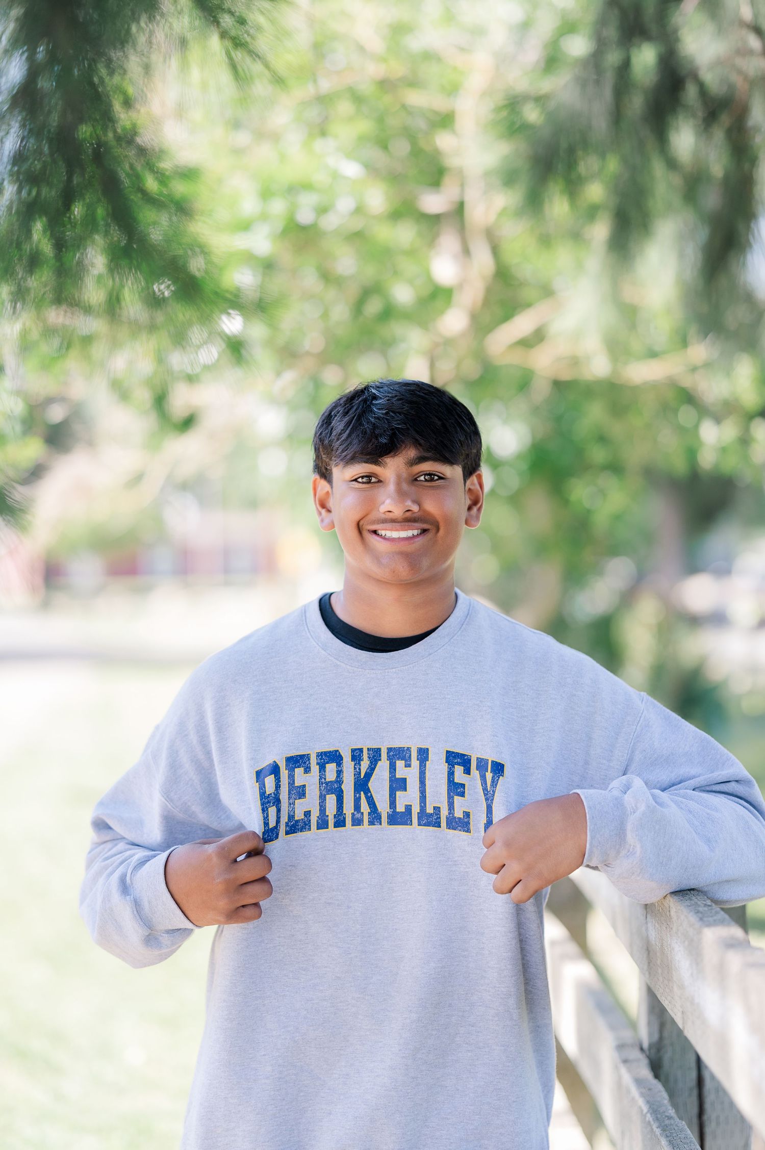 Smiling young man wearing a gray Berkeley crewneck sweatshirt, standing outdoors near a wooden fence.
