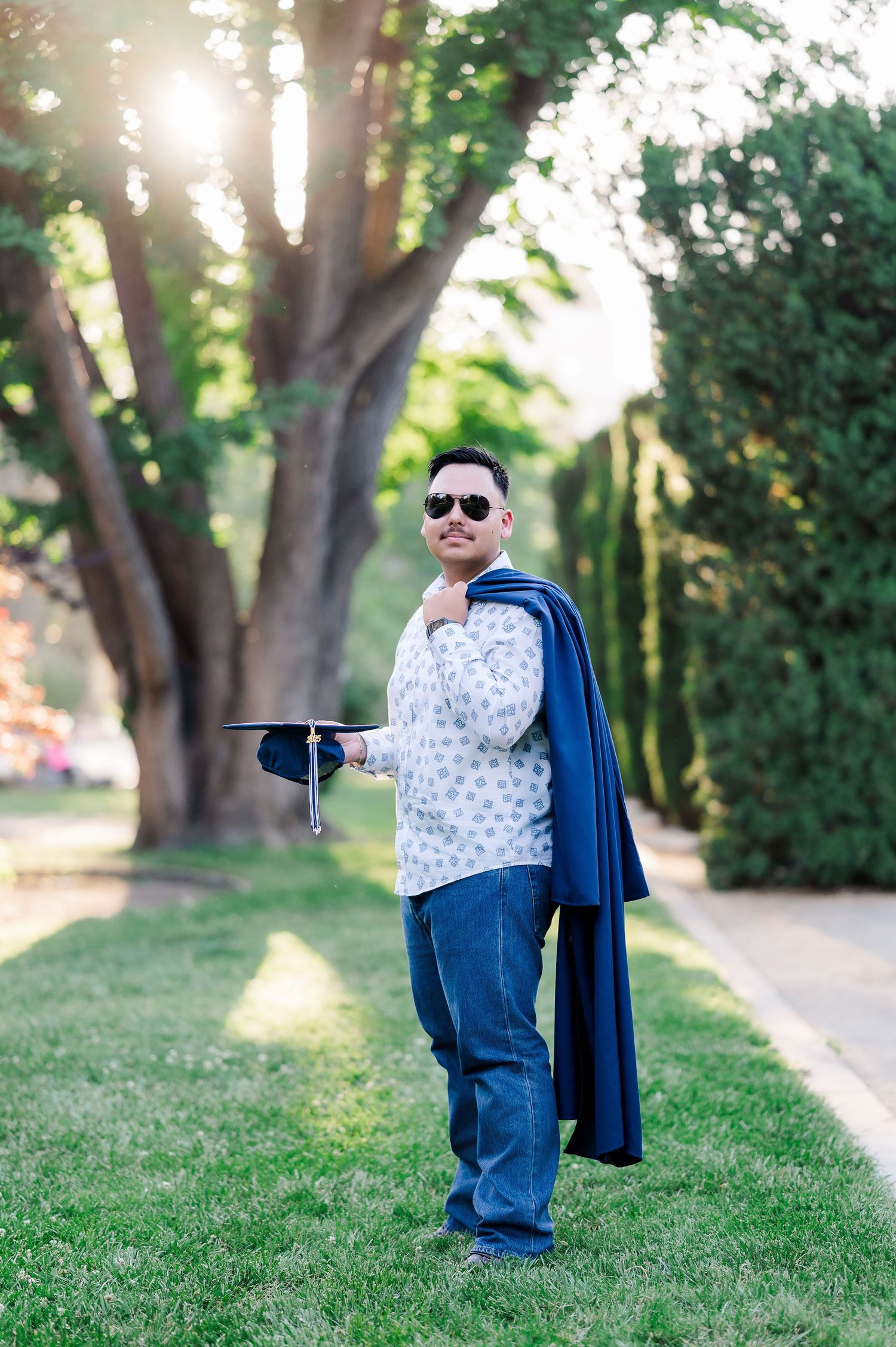 Man in sunglasses holding graduation cap and gown outdoors on green lawn with trees in background.