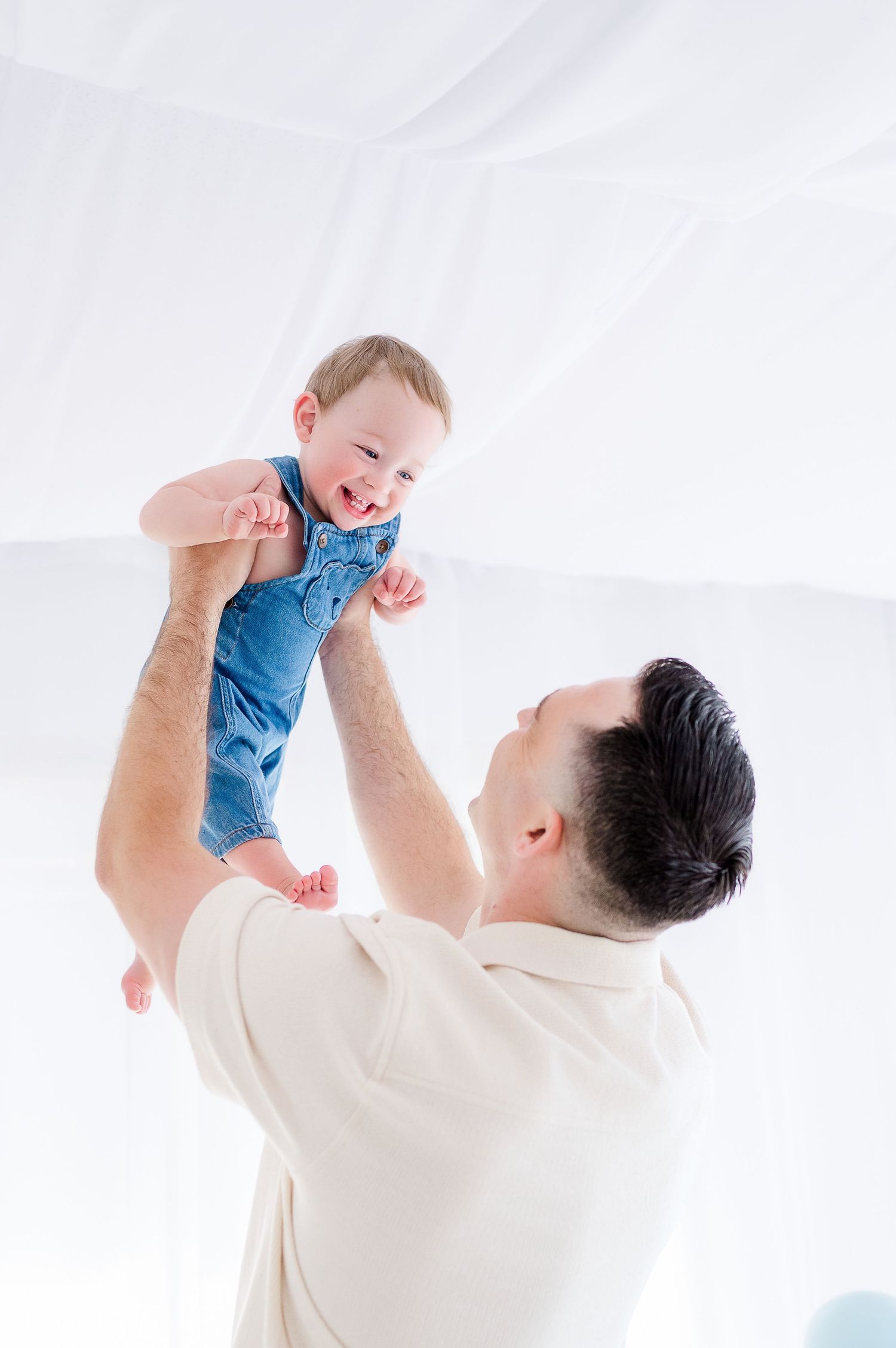 A happy father lifts his smiling baby in a blue onesie high in the air in a bright white room.