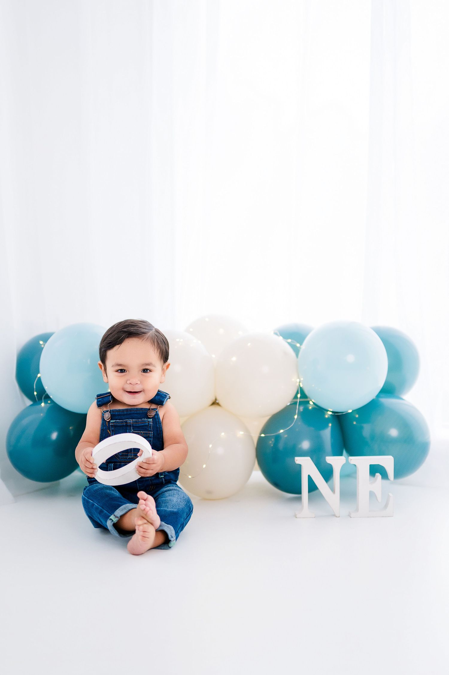 A smiling baby in denim overalls sits among teal and white balloons with letter decorations spelling ONE for a first birthday.