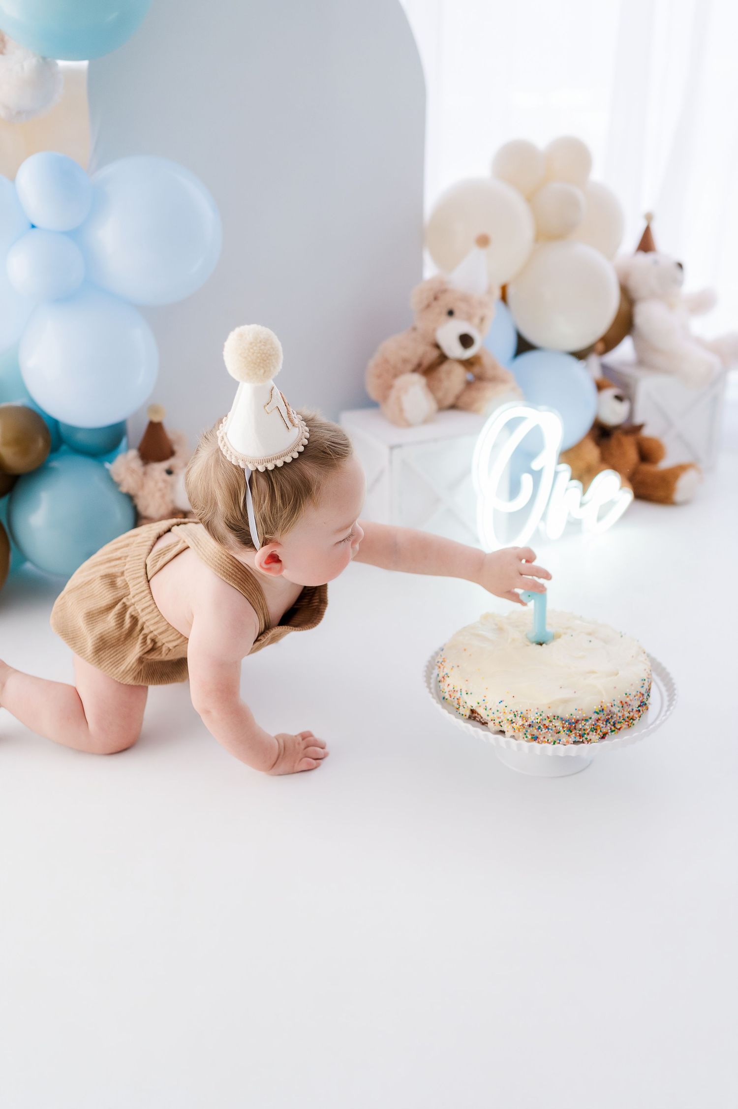 Baby in party hat reaches for first birthday cake surrounded by blue balloons and teddy bears at a smash cake celebration.
