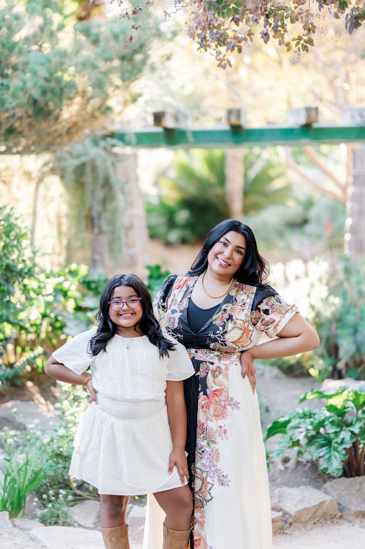 Mother and daughter wearing matching white dresses pose playfully in a garden setting with natural lighting.