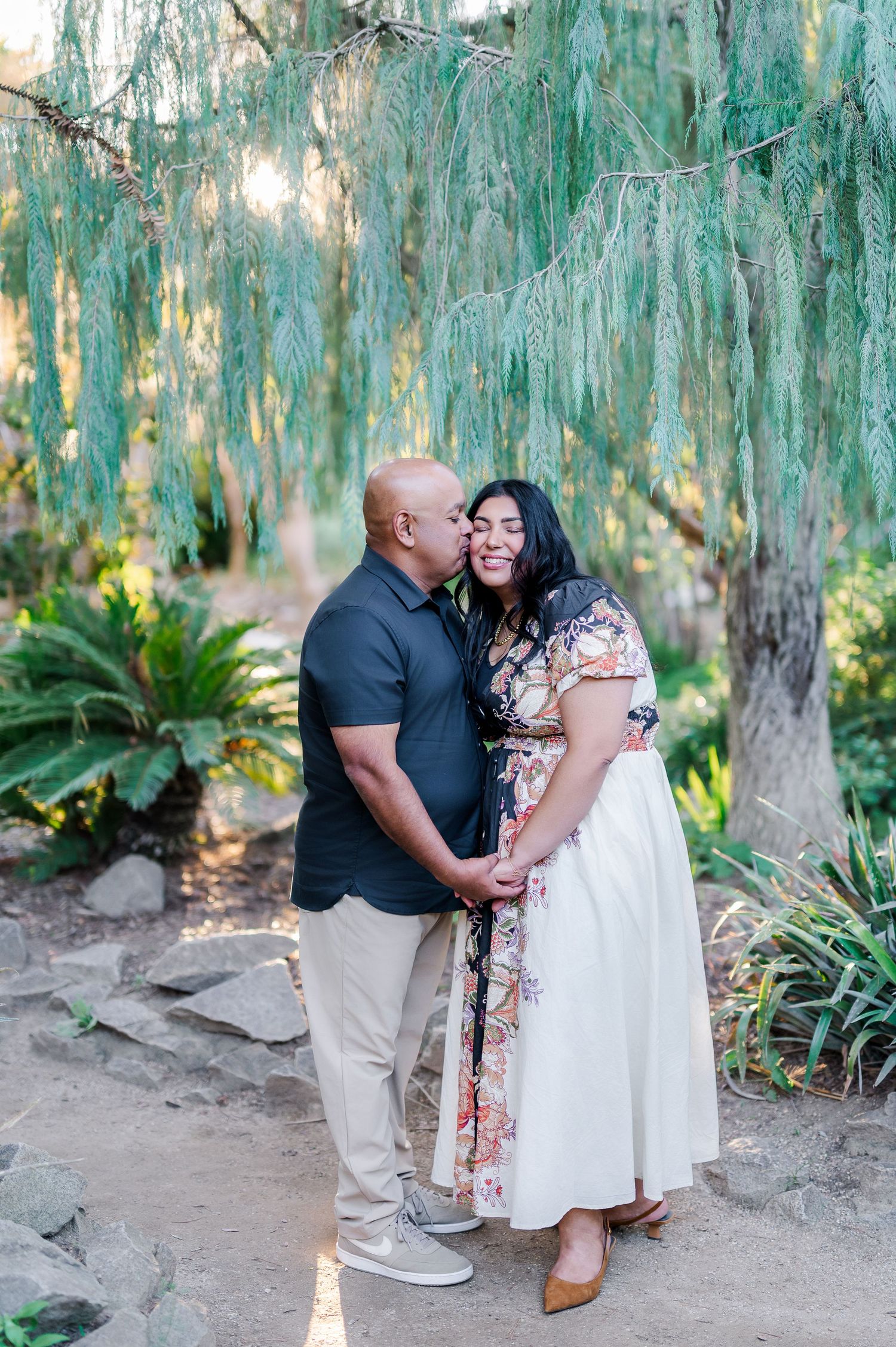A romantic couple embraces under cascading willow trees in a garden setting with soft natural lighting.