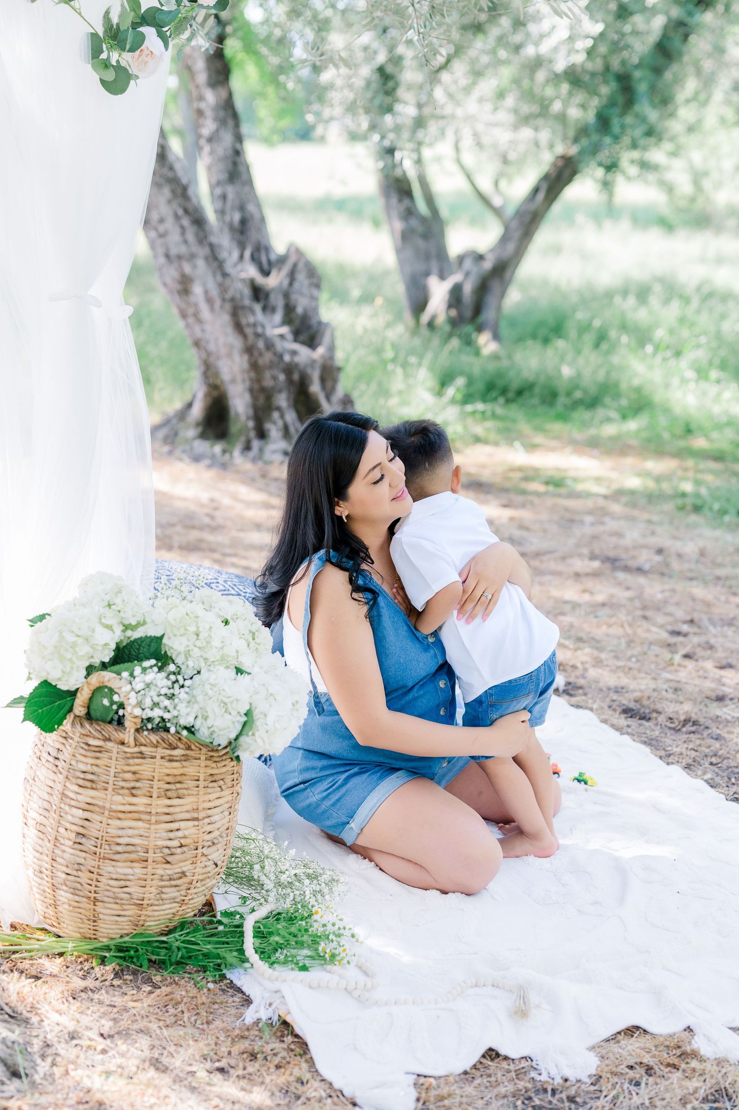 Couple sharing intimate moment on white blanket in olive grove with wicker basket and flowers nearby.