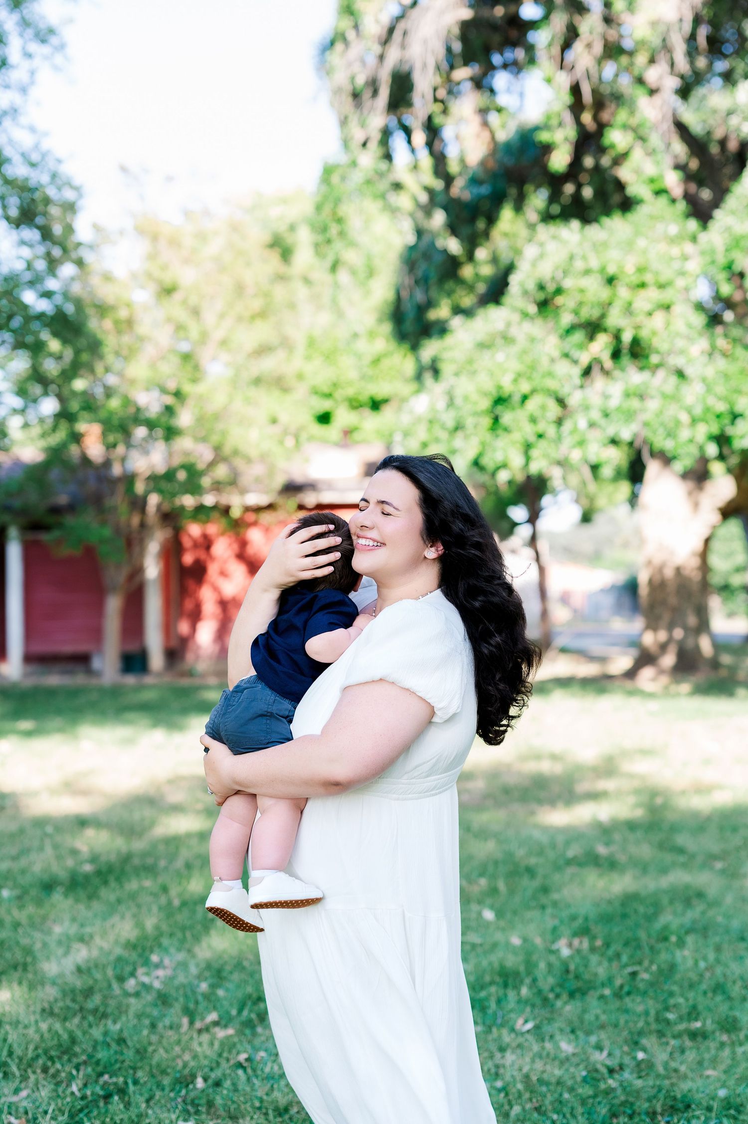 A person in white dress joyfully holds a small child in a sunny backyard setting.