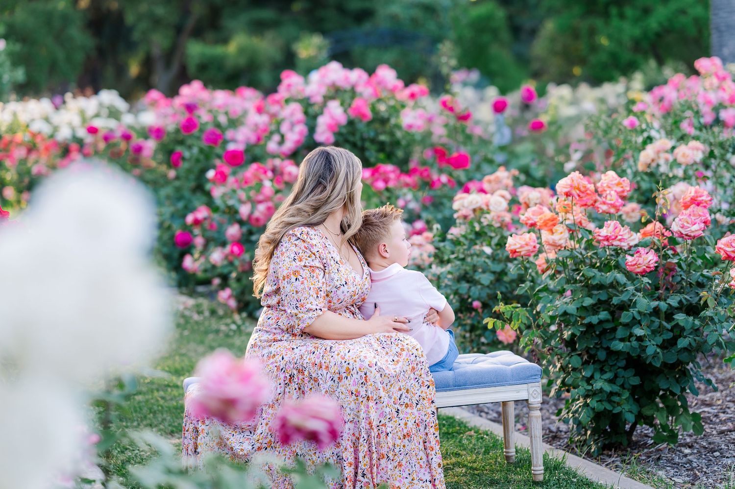 A mother and child share a tender moment on a bench surrounded by pink roses.
