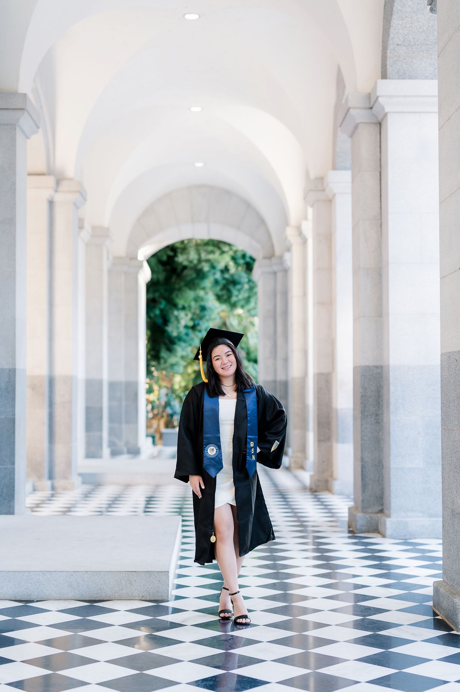 Graduate in black gown posing playfully in elegant colonnade with checkered floor.