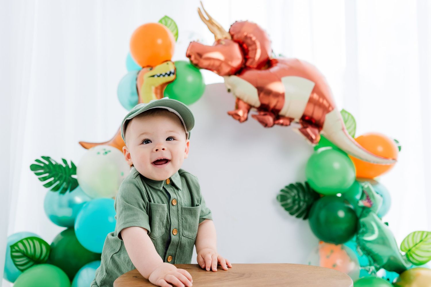 Baby boy in green outfit smiles at dinosaur-themed first birthday party with colorful balloon arch backdrop.