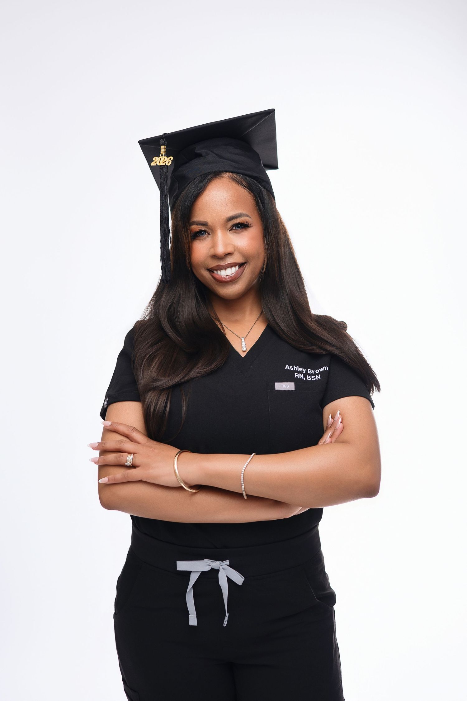 Smiling woman in black scrubs and graduation cap poses with arms crossed against white background.