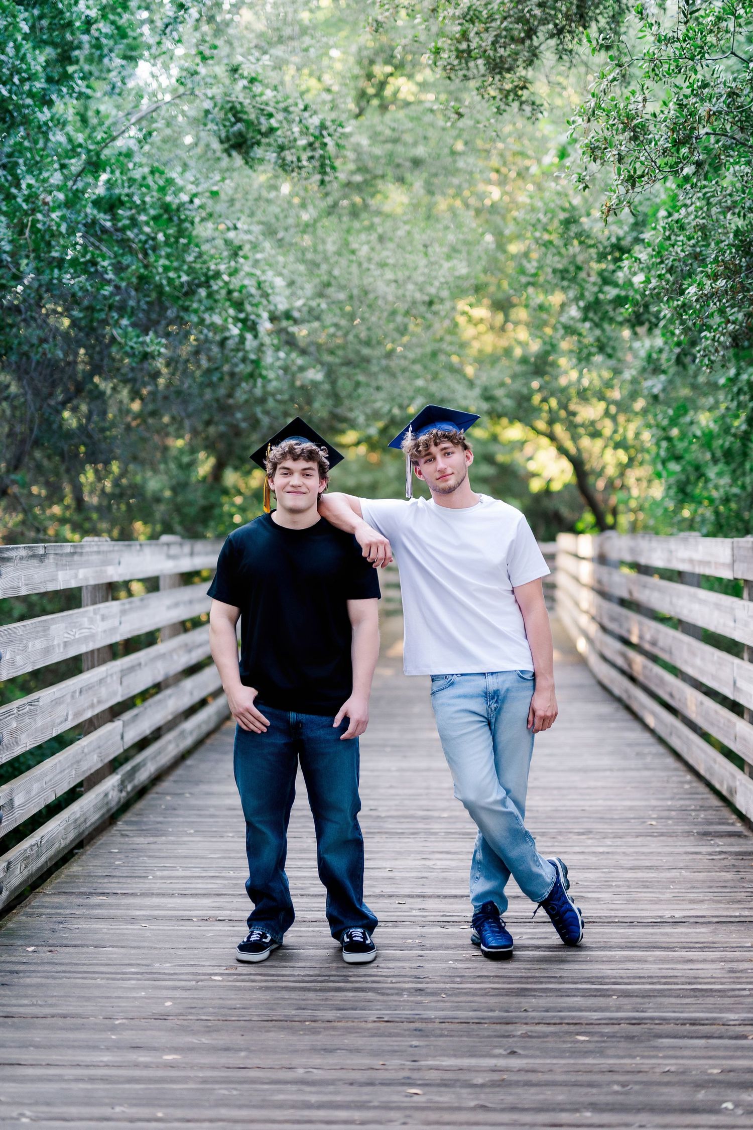 Two young men in graduation caps pose on a wooden bridge surrounded by lush green trees.