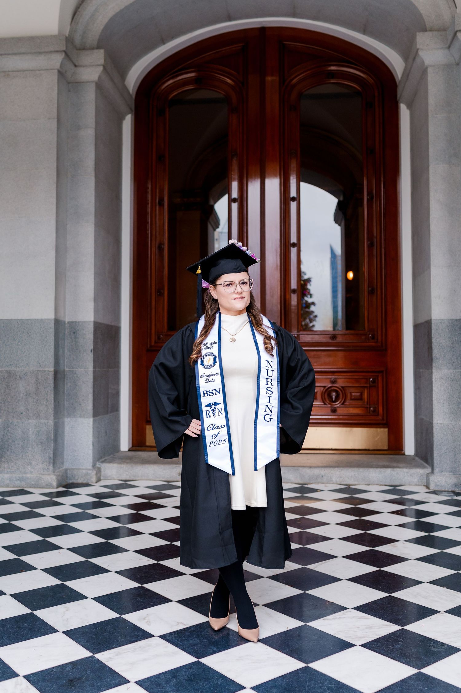 Graduate in black cap and gown with honor stole stands on checkered floor before ornate wooden doors.
