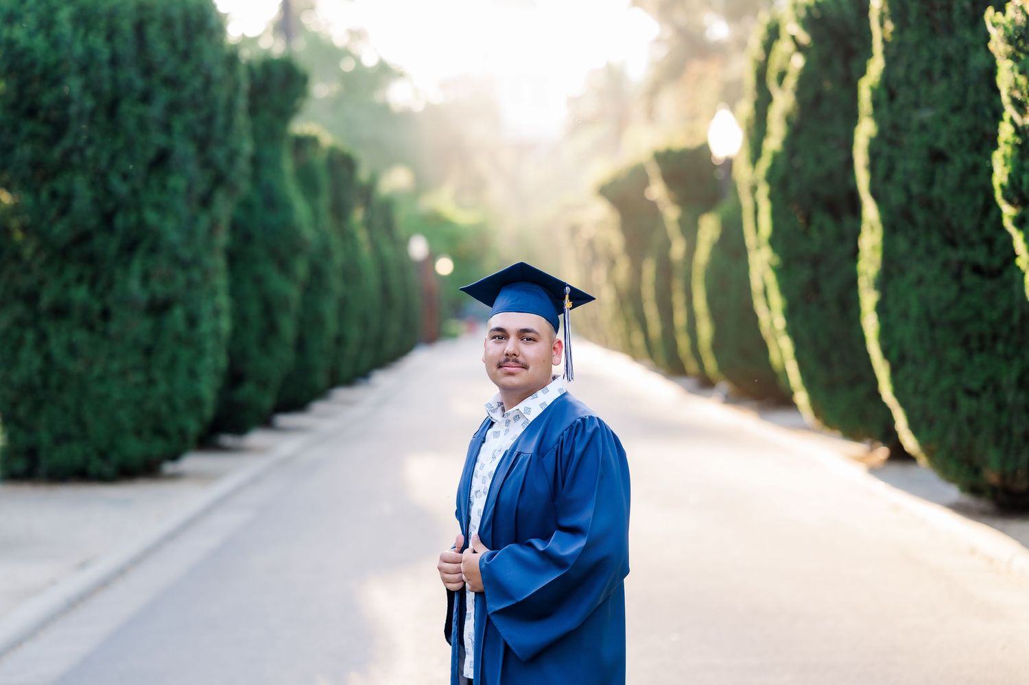 A graduate in blue cap and gown poses on a cypress-lined path at sunset.