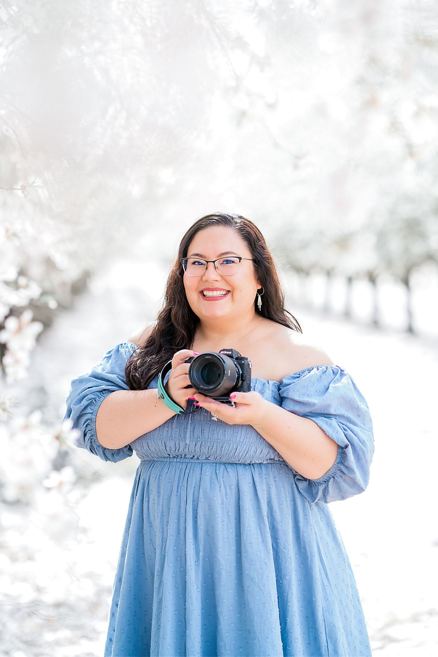 Photographer in blue off-shoulder dress holds camera in snowy orchard setting.