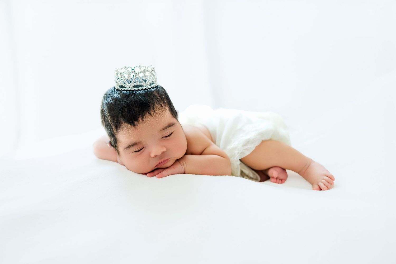 A sleeping newborn wearing a tiny crown lies peacefully on white fabric.