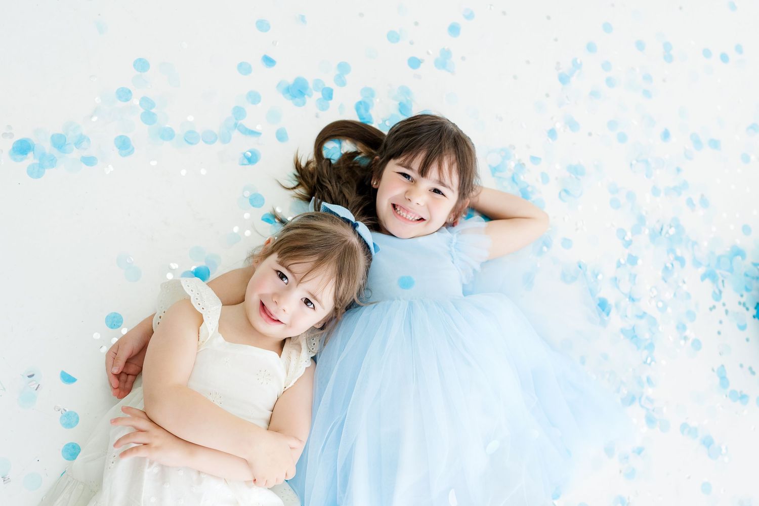 Two smiling girls in white and blue dresses lying on a white floor surrounded by blue confetti petals.