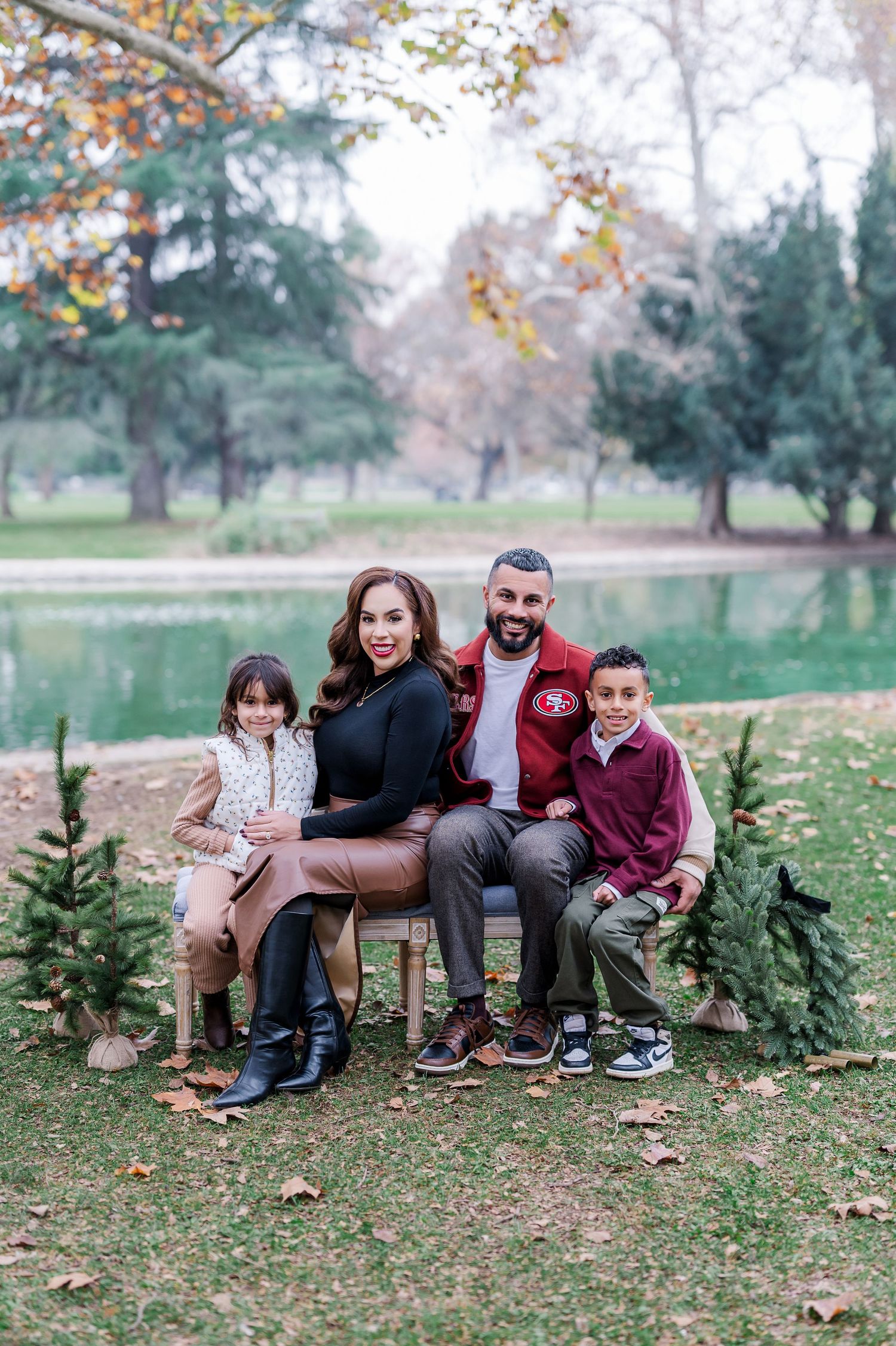 Family of four posing outdoors near a pond with small Christmas trees, wearing casual fall attire.