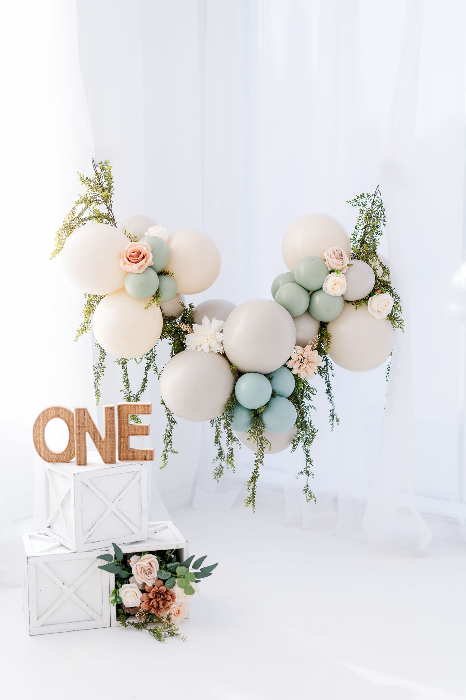 First birthday party decoration with rose gold 'ONE' sign, pastel balloon arch and floral arrangements against white backdrop.