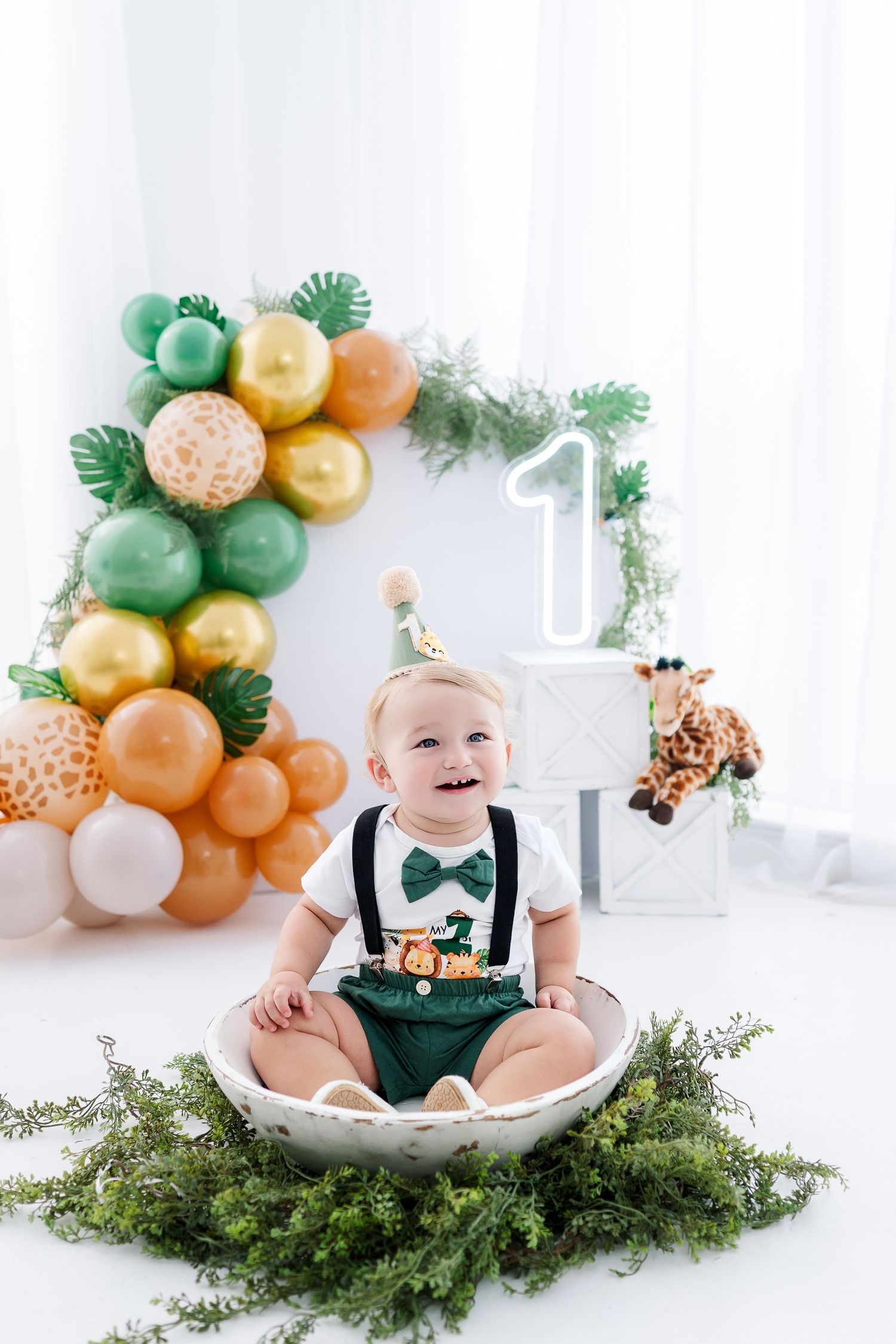 Safari themed first birthday cake smash photo session with green and gold balloon arch and tropical decor.