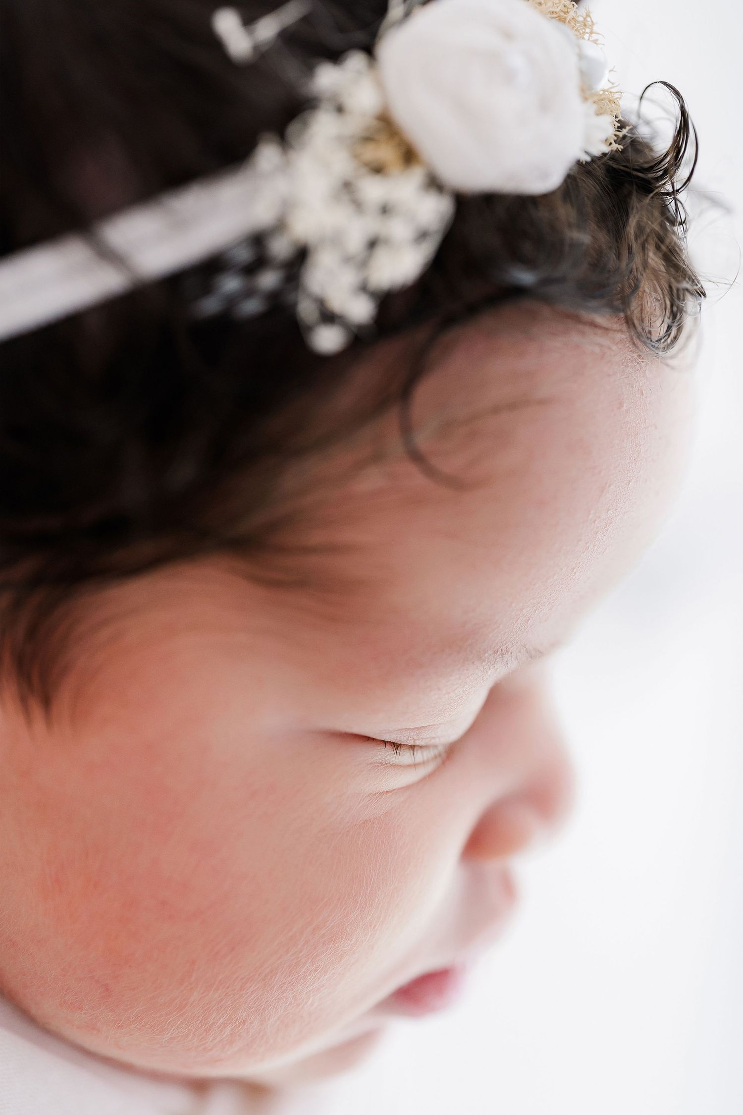 Close-up view of a sleeping baby wearing delicate white floral hair accessories against a soft white background.