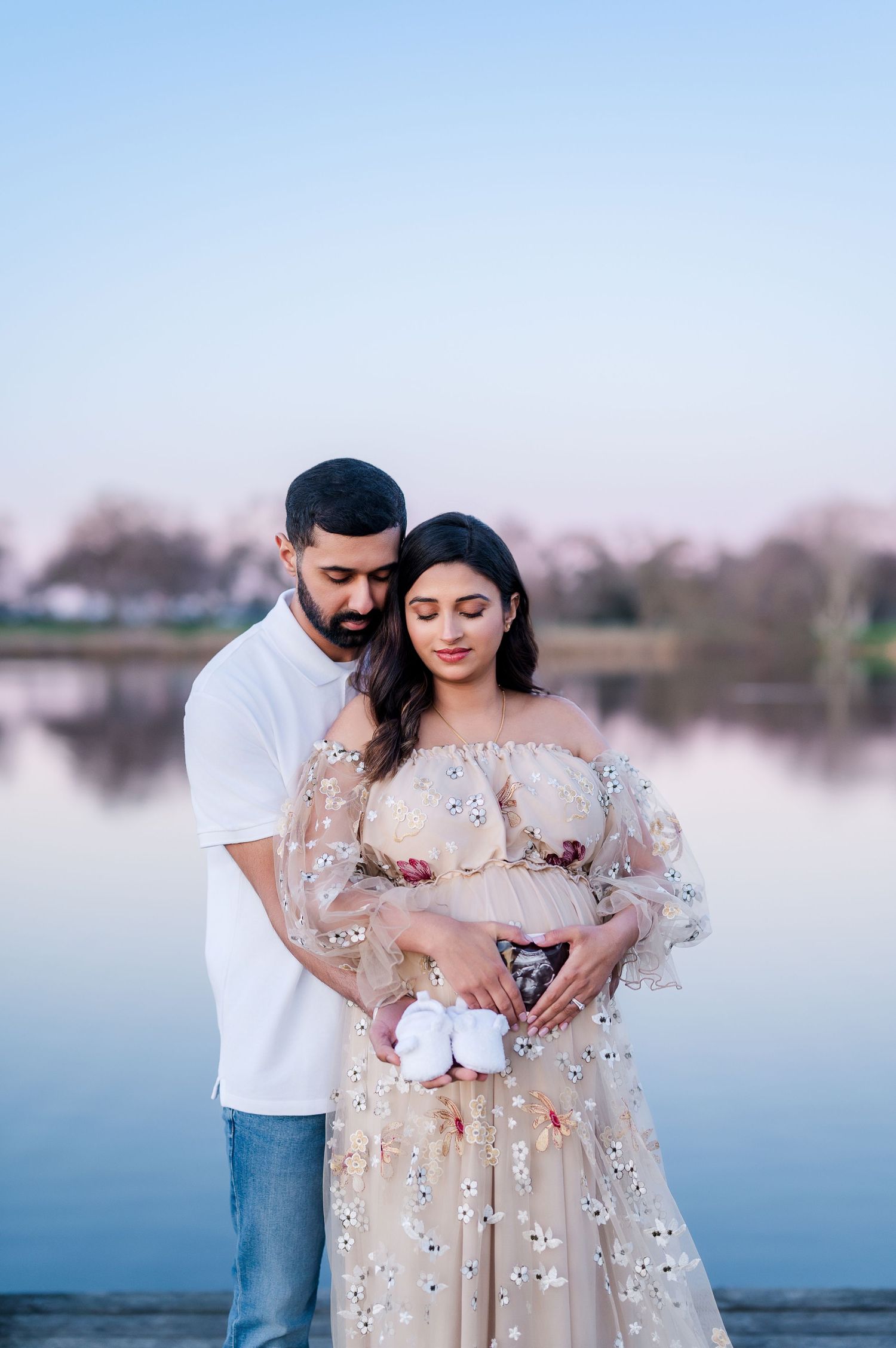 A couple in romantic maternity photoshoot wearing white and floral dress poses by a serene lake at sunset.