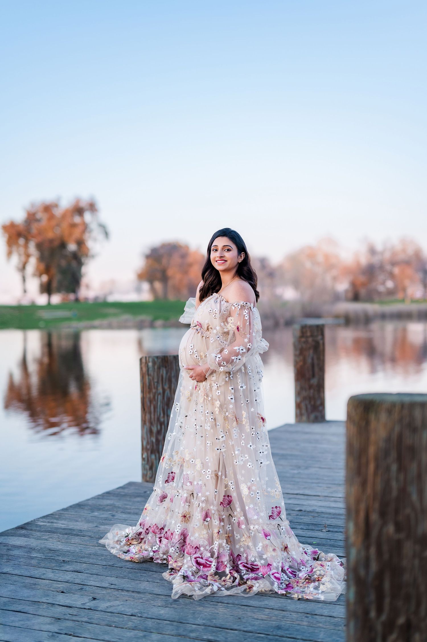 A maternity photo shoot on a lakeside dock featuring a white lace dress with purple floral details.