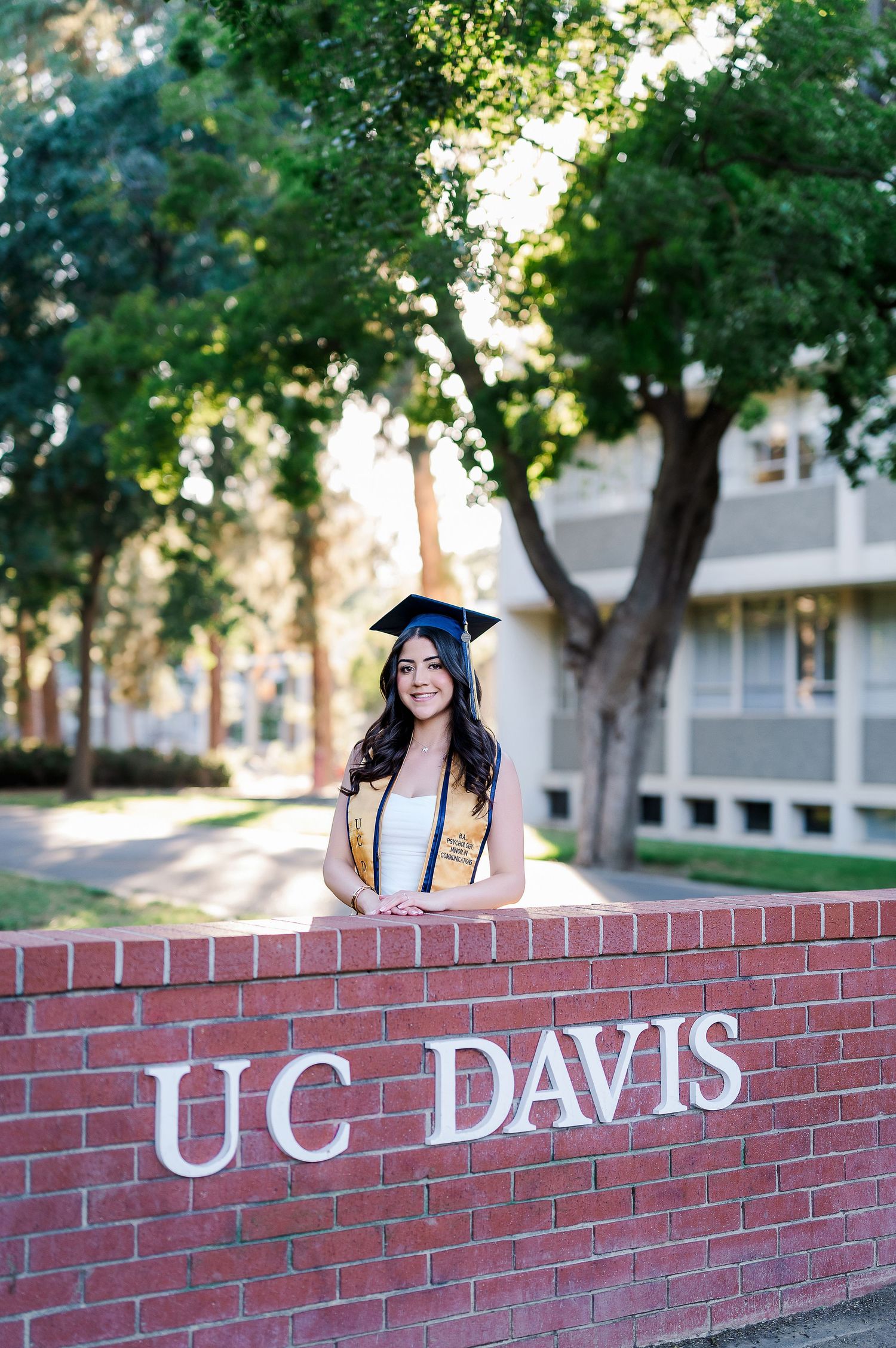 Graduate in cap and gown poses on UC Davis brick sign wall during golden hour.