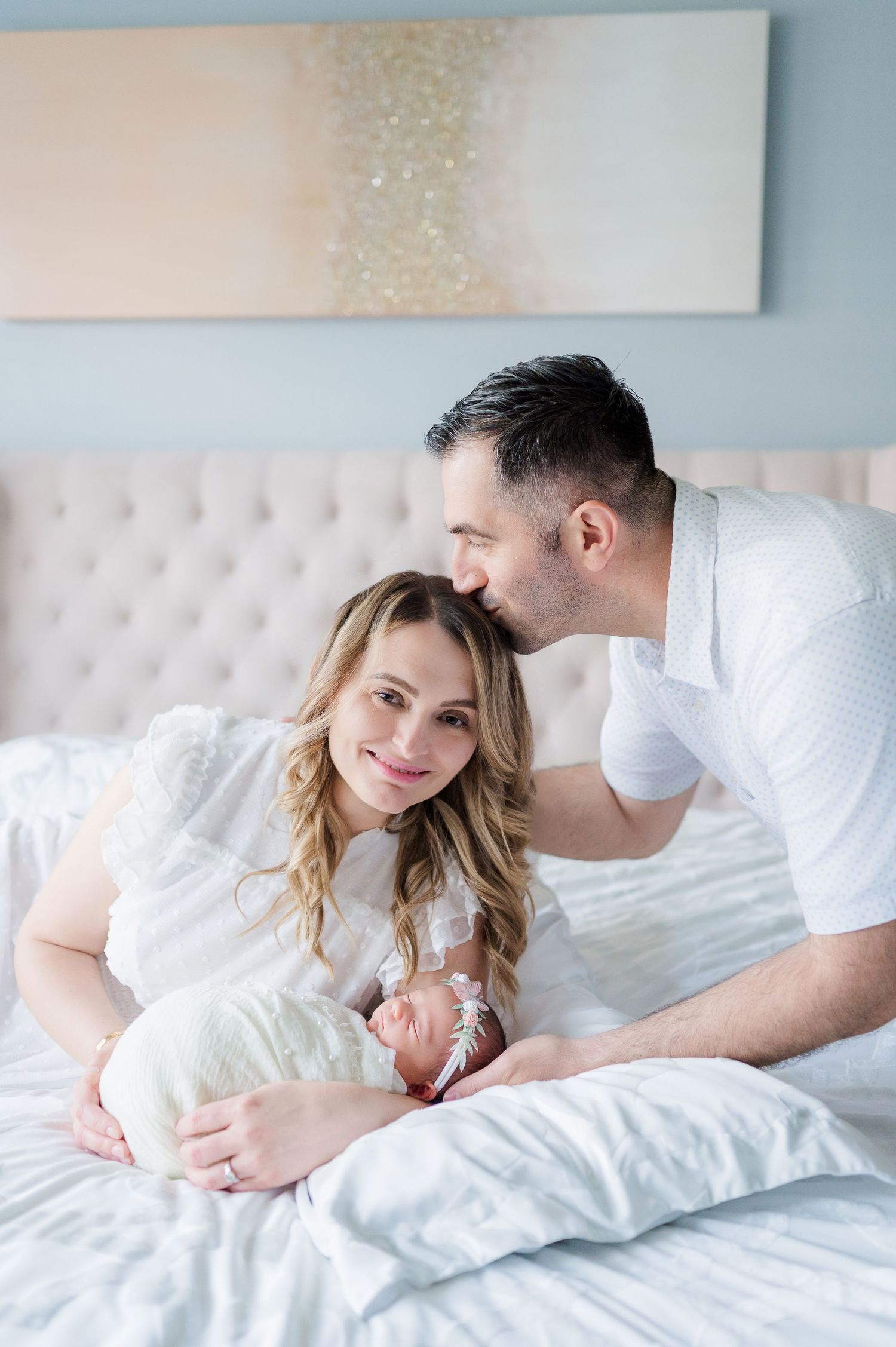 Intimate bedroom portrait featuring a newborn baby swaddled in white with parents on cozy bedding.