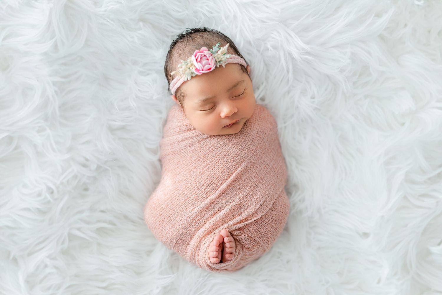Newborn baby wrapped in pink swaddle with floral headband sleeping on white fluffy rug.