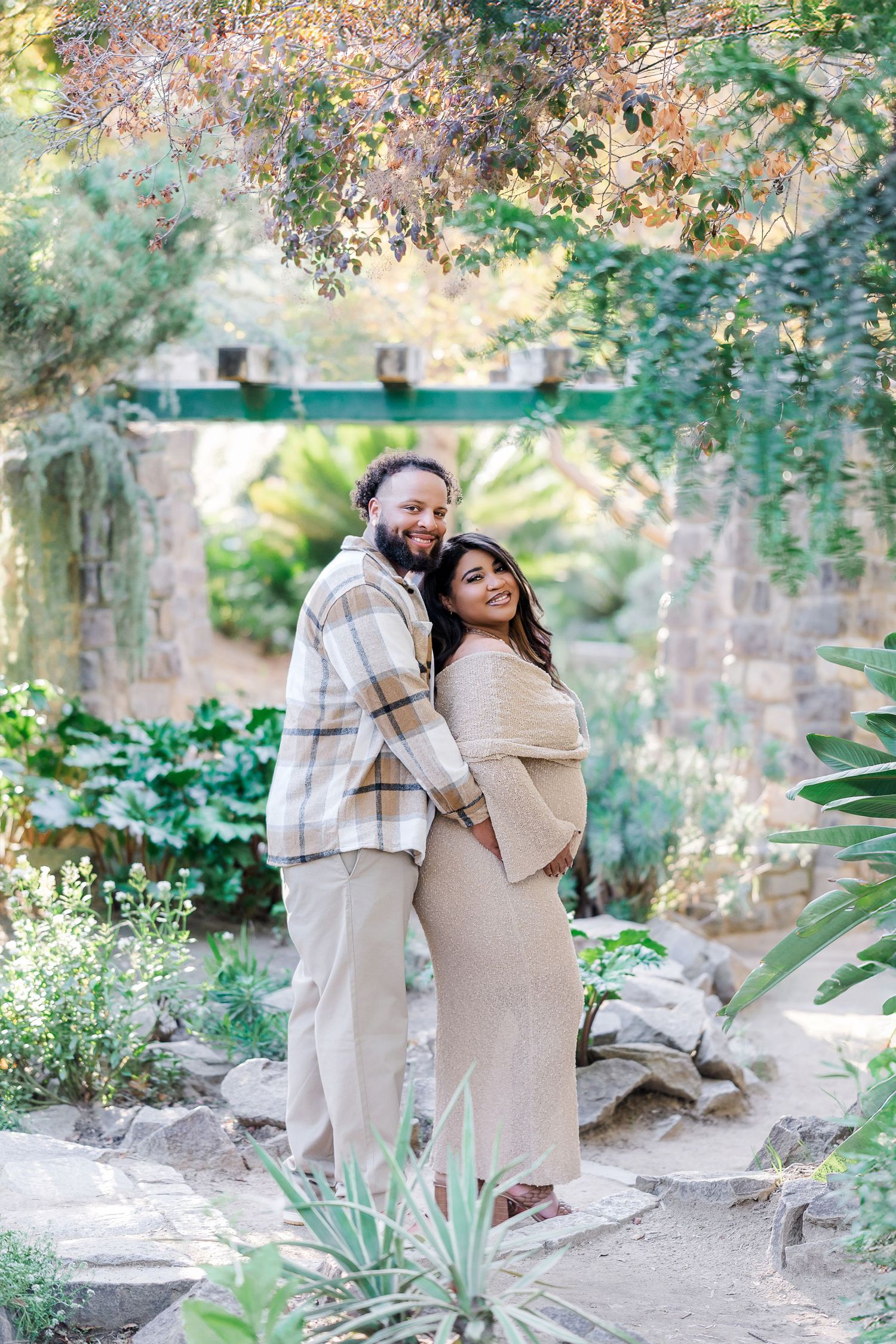 A couple embraces tenderly in a garden setting with autumn foliage and stone walls creating a romantic atmosphere.