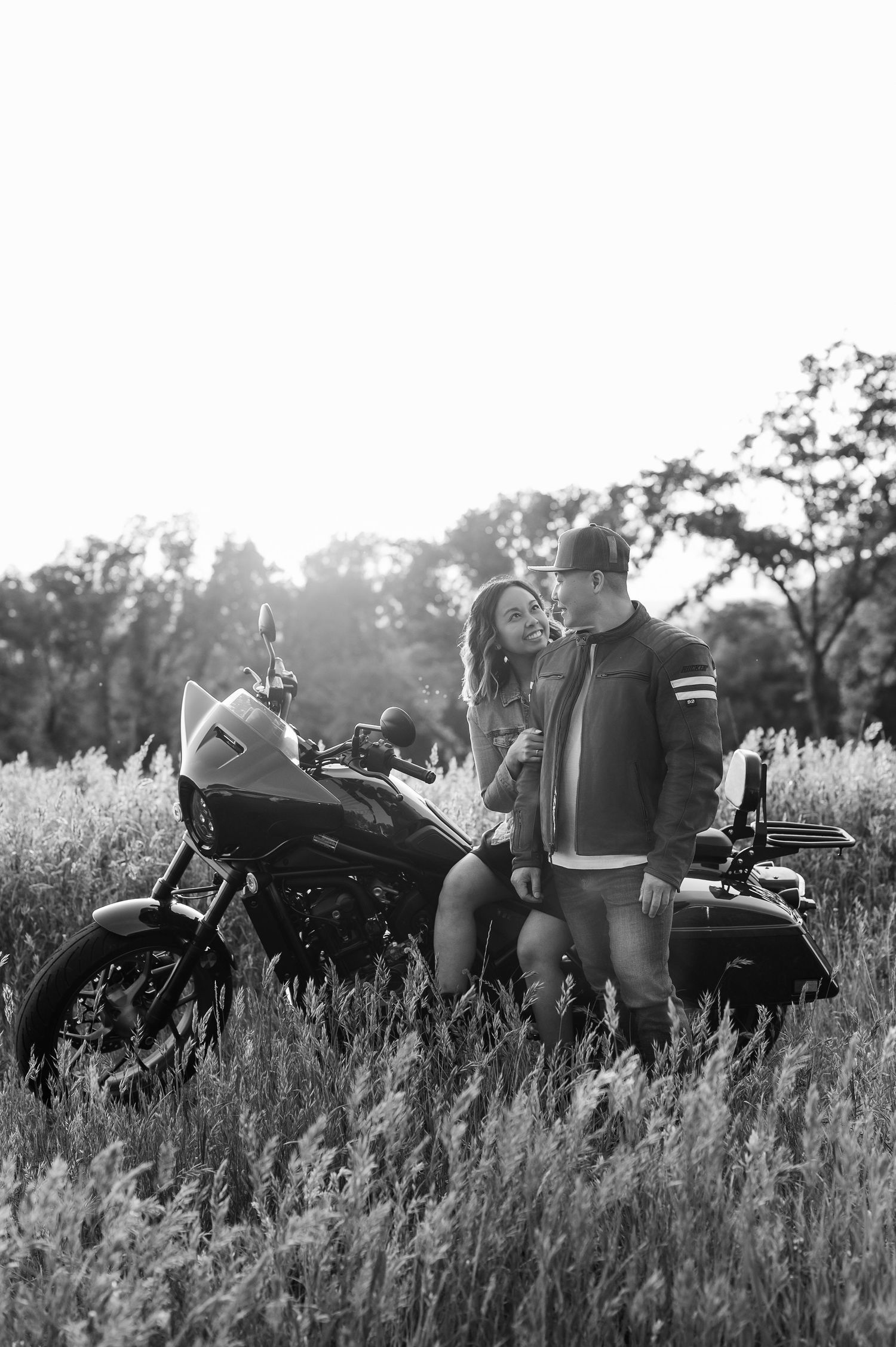 Romantic black and white photo of couple with motorcycle in tall grass at sunset.