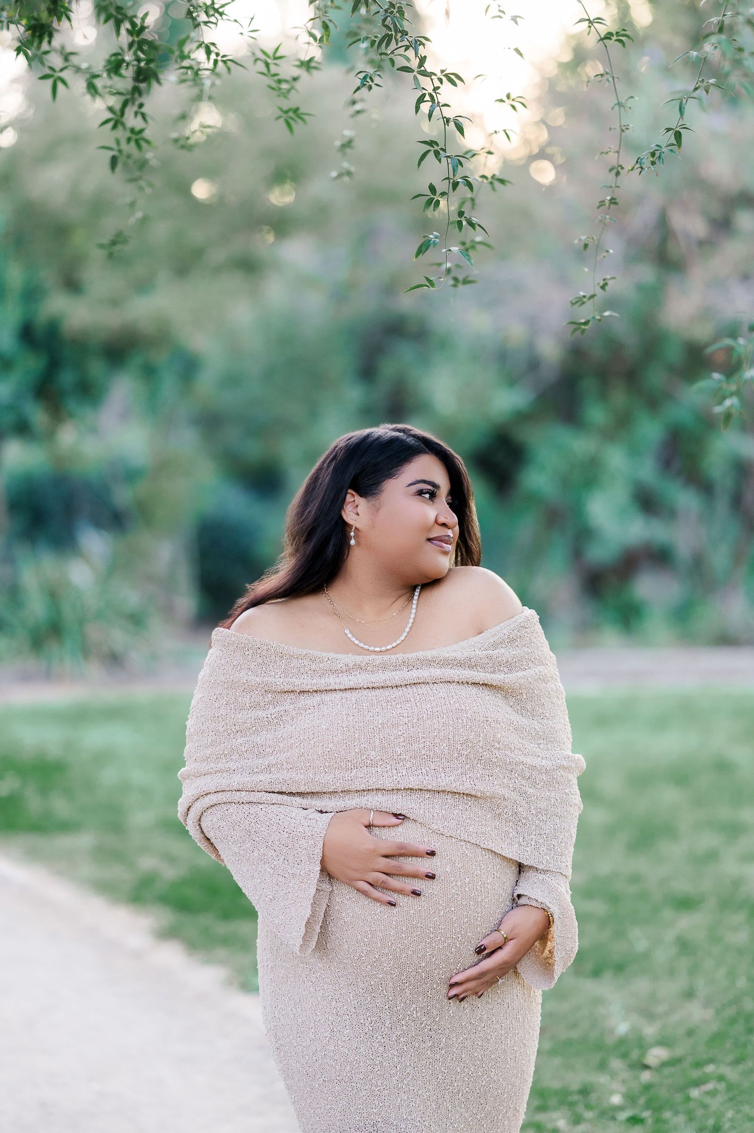 Expectant figure wrapped in beige shawl poses outdoors with green foliage in soft bokeh background.