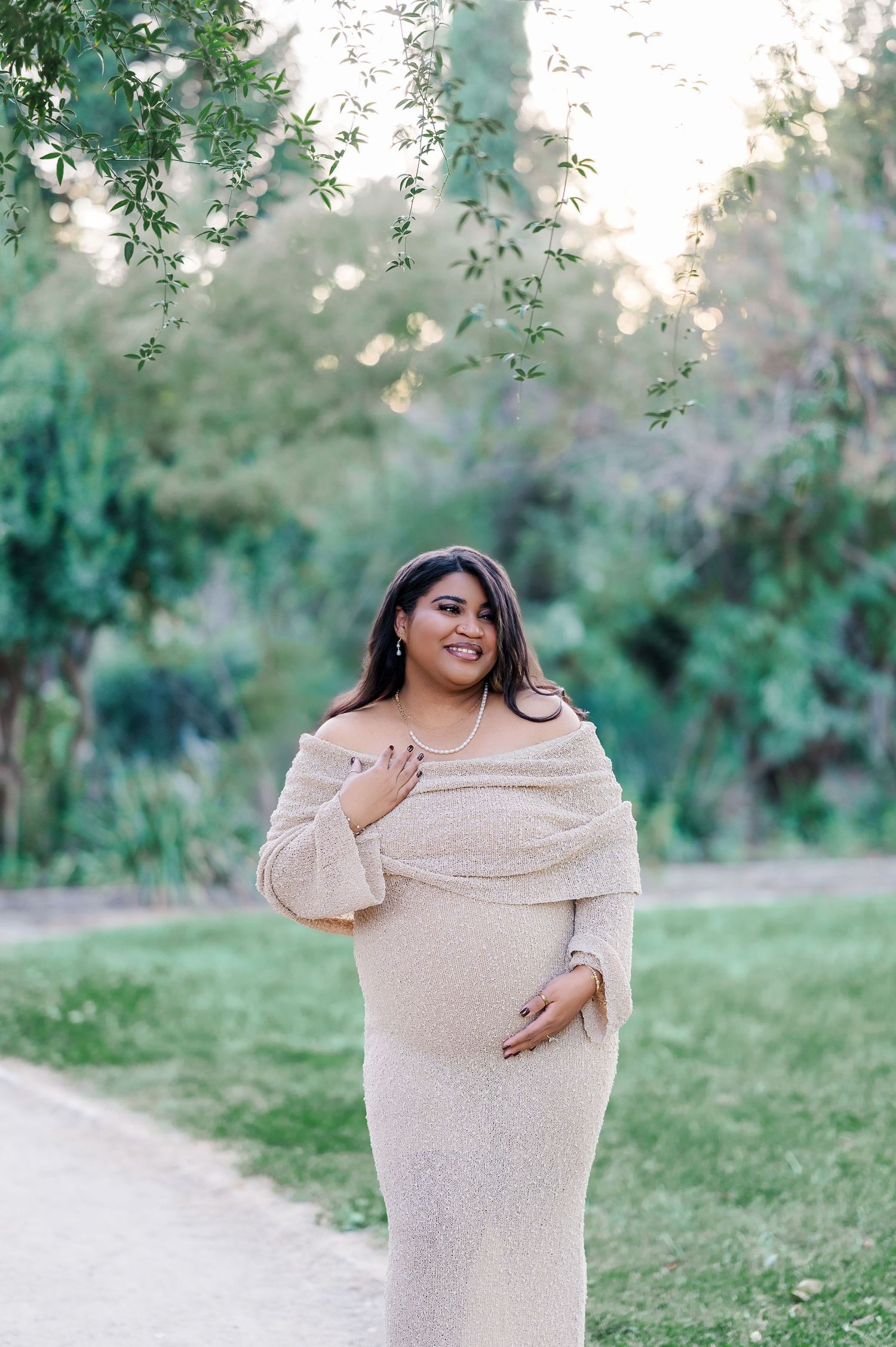 Woman in beige sweater poses joyfully outdoors in a natural setting with green foliage in the background.