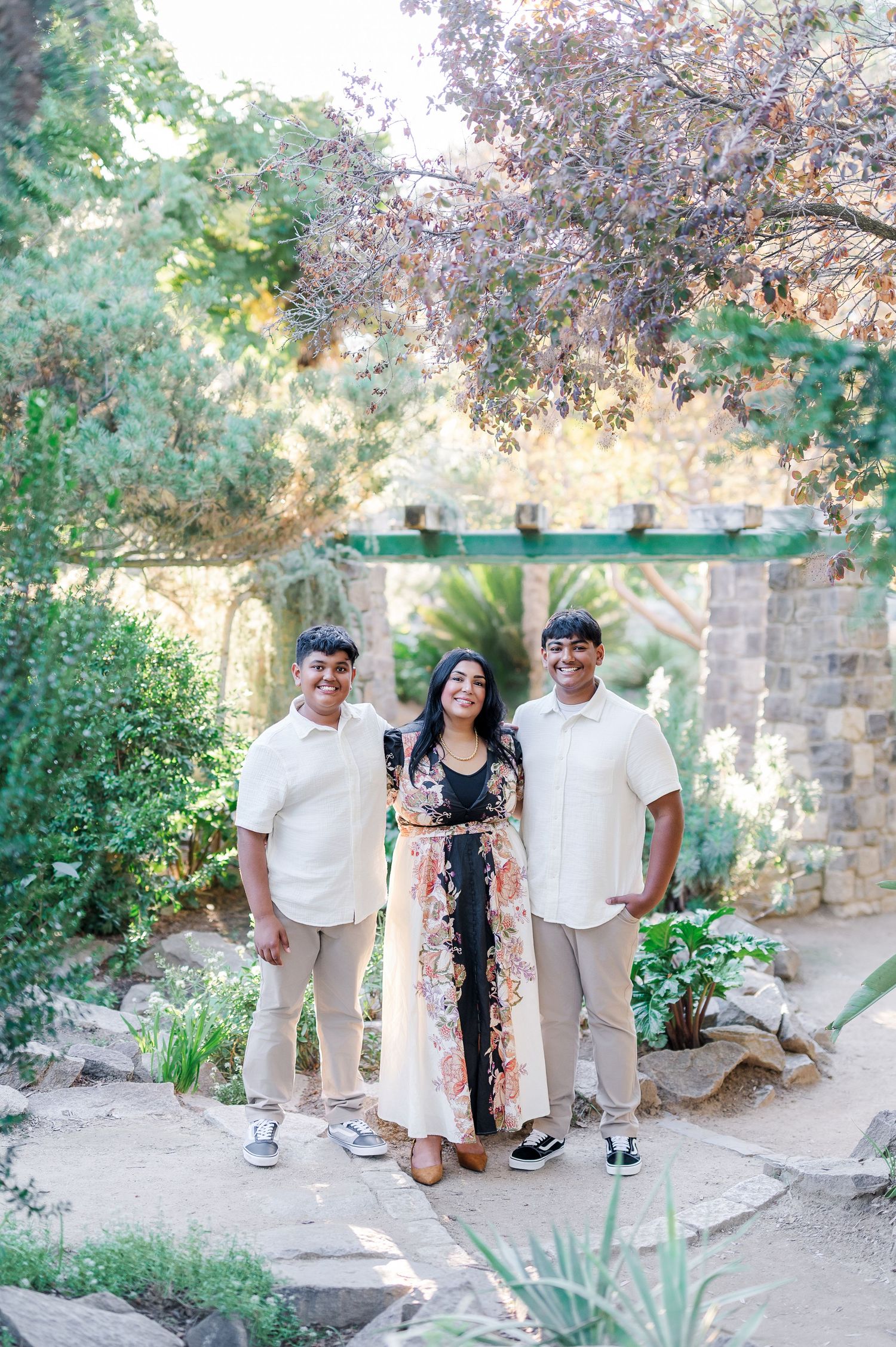 Three people in coordinated white and black outfits pose together in a lush garden setting during a photo session.