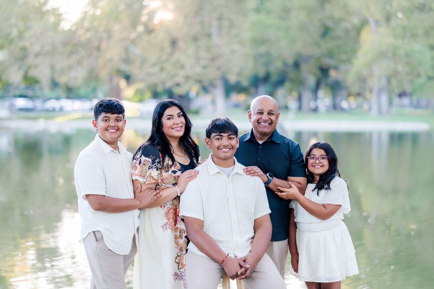 A family group poses together wearing coordinated white outfits near a serene lake during a sunny outdoor photo session.