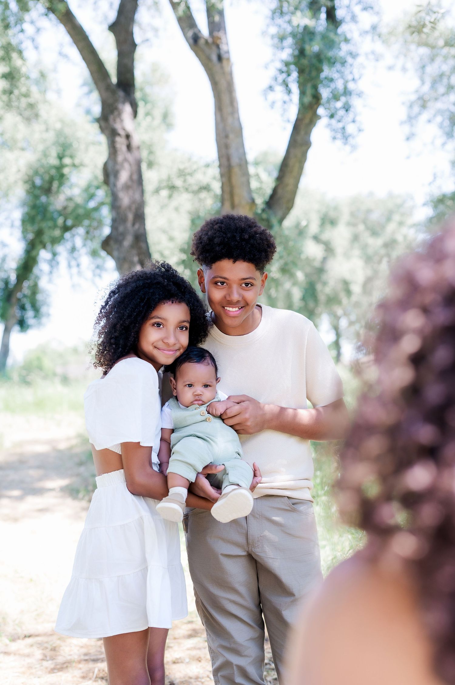 Family wearing neutral colors poses together in a sunny outdoor setting with trees in the background.