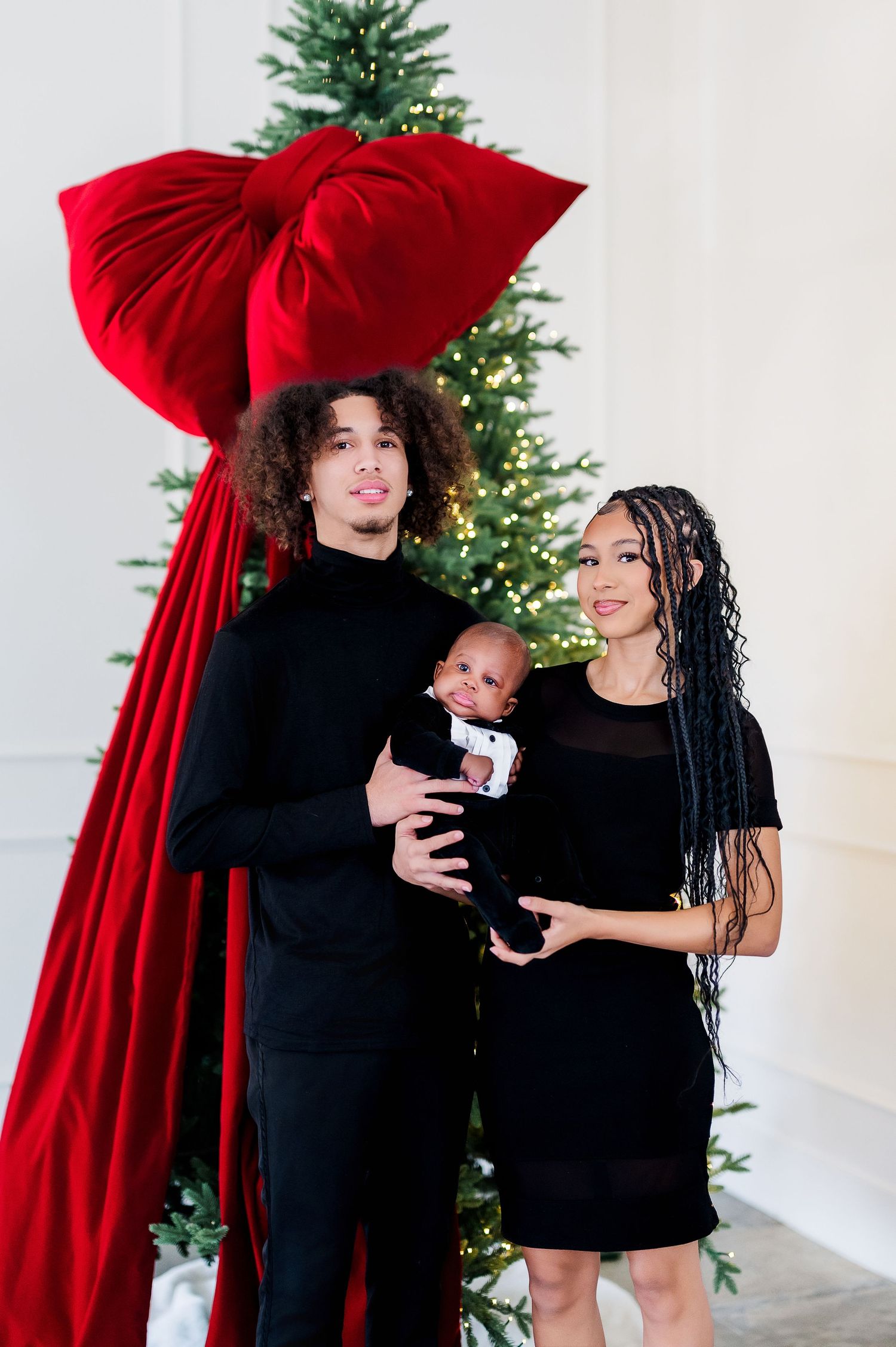 Family poses in front of Christmas tree with giant red bow backdrop for holiday portrait.