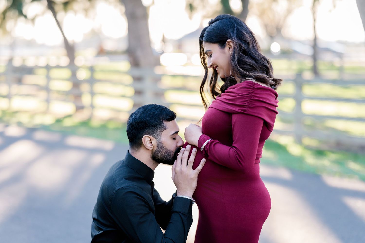Intimate maternity moment captured between couple in burgundy dress against wooded background.